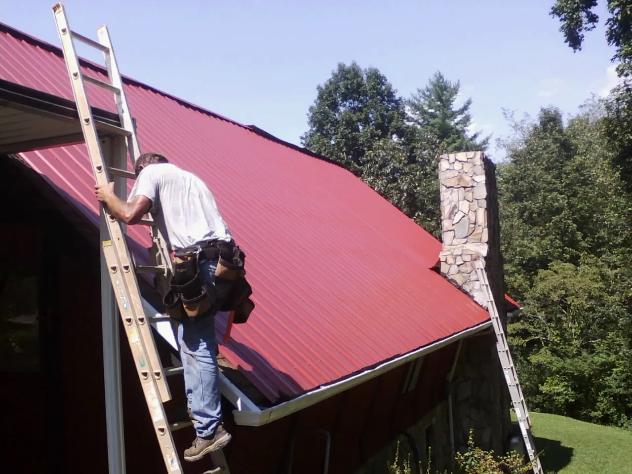 Roofer on a ladder beside a red metal roof and stone chimney — Soffit & Fascia Repair by Roofing Brothers in West Newfield
